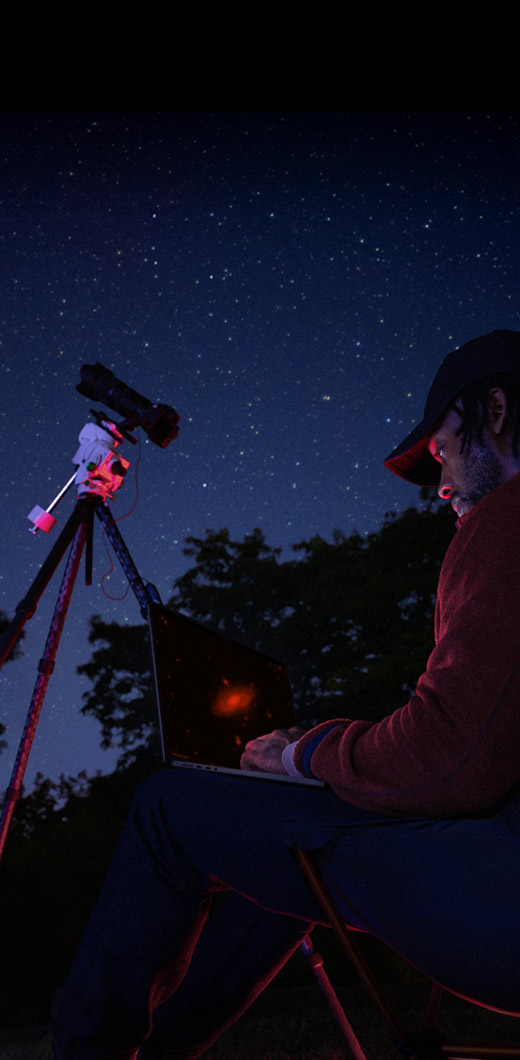 夜に屋外でMacBook Proを膝の上に置いて使っている人物。MacBook Proは星空に向けられた三脚の上のデバイスに接続されている