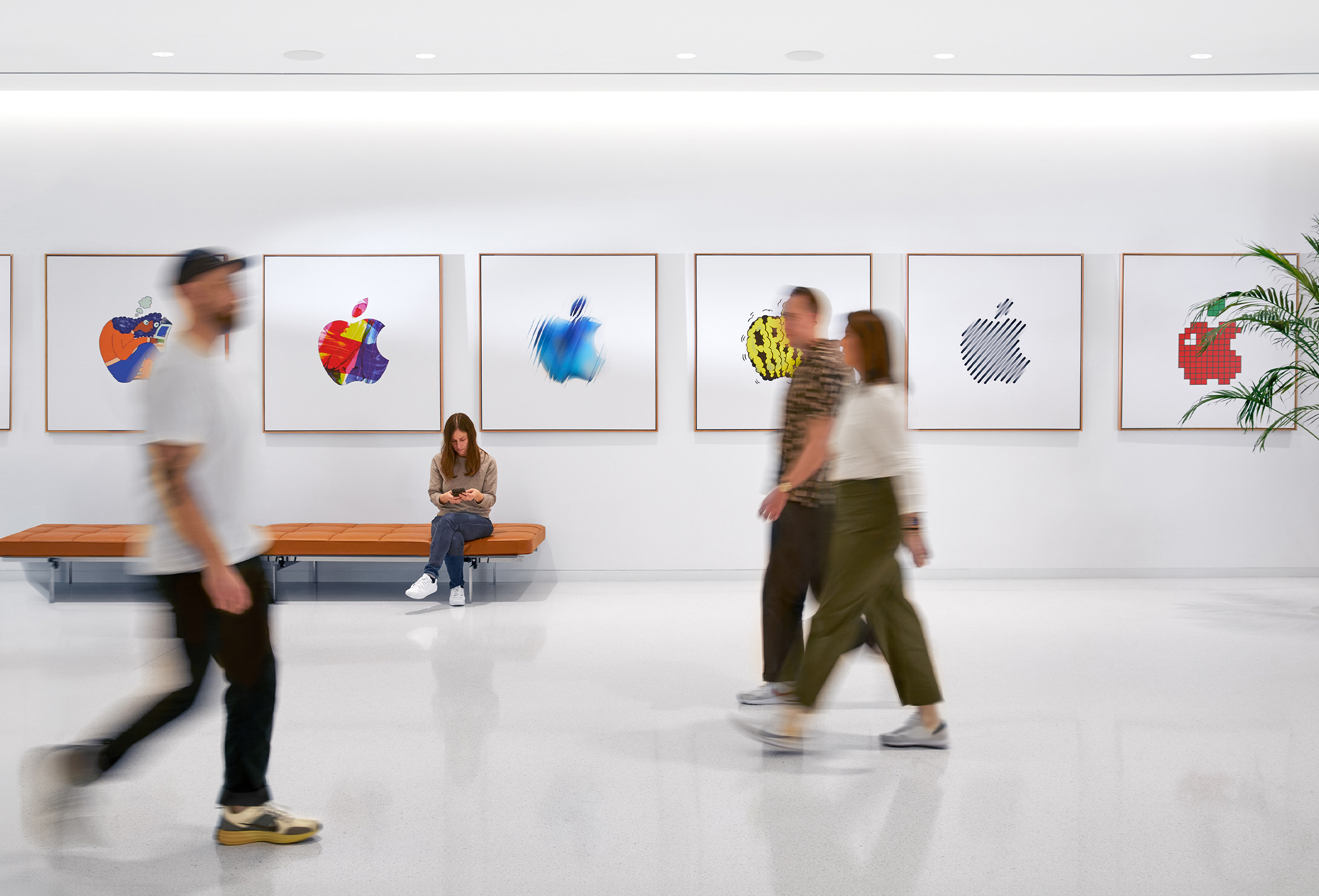 三位 Apple 同事走過有著色彩繽紛 Apple 標(biāo)誌的室內(nèi)牆面，第四位員工坐在長椅上。