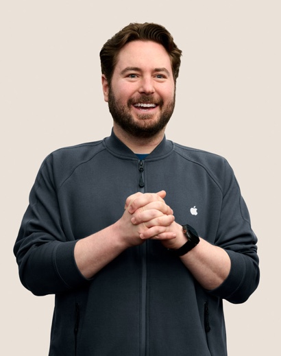 Portr?t eines l?chelnden Apple Retail Teammitglieds mit kurzen, gewellten dunklen Haaren und Bart.