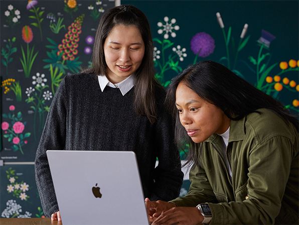Twee teamleden van Apple Marketing die samenwerken op een MacBook. Ze zitten voor posters die hun team heeft ontworpen.
