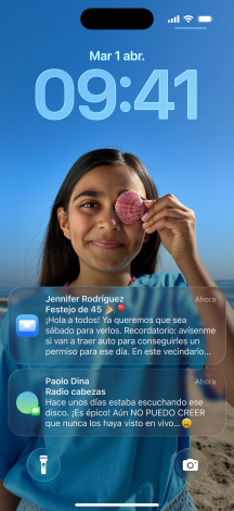 Cinco modelos de iPhone se separan uno detrás del otro, cada modelo se superpone levemente sobre el siguiente, la pantalla del modelo ubicado más a la izquierda muestra una conversación con un fondo en Mensajes, el siguiente muestra la pantalla de inicio con íconos de apps coloridos, el del centro muestra la pantalla bloqueada con una foto y los números de la hora dise?ados con Liquid Glass, así como dos notificaciones en la parte inferior de la pantalla, el siguiente modelo hacia la derecha muestra íconos de apps translúcidos en Liquid Glass y el último a la derecha muestra el reproductor de música hecho con Liquid Glass