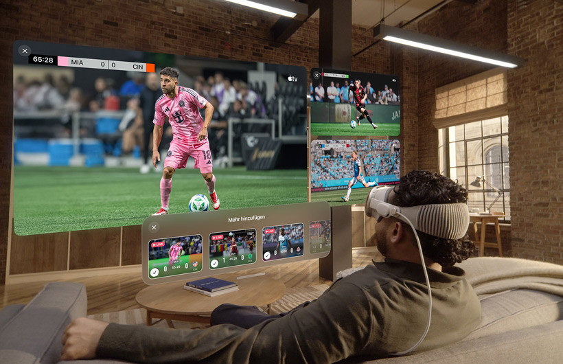 Eine Person mit einer Apple Vision Pro sitzt auf einem Sofa in einem Wohnzimmer. Vor ihr wird ein gro?er Videobildschirm neben zwei kleineren Bildschirmen gezeigt, die jeweils ein anderes Sportereignis zeigen. Unter dem gro?en Bildschirm befindet sich ein Auswahlfenster mit dem Text ?Mehr hinzufügen“