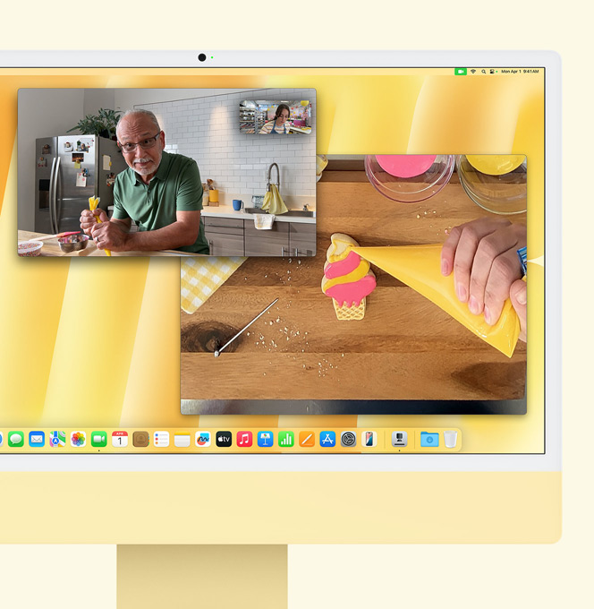 Ein iMac in Gelb. Eine Demonstration der Schreibtischansicht. Auf dem Bildschirm wird eine B?ckerin in einem Videoanruf mit einer anderen Person gezeigt. Auf einem Bildschirm werden die B?ckerin und das Gesicht des Mannes gezeigt. Auf dem anderen Bildschirm werden die H?nde der B?ckerin beim Glasieren eines Kekses gezeigt