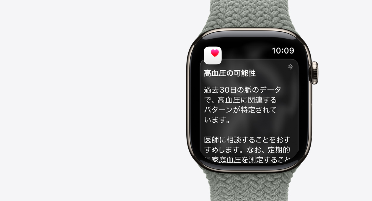 ナチュラルチタニウムケースのApple Watch Series 11。高血圧の兆候を示す通知と、ピンクのハートマークが入った白いボックスが表示されている。右側(cè)面の外観にはDigital Crownがあり、グリーングレイのブレイデッドソロループが付いている