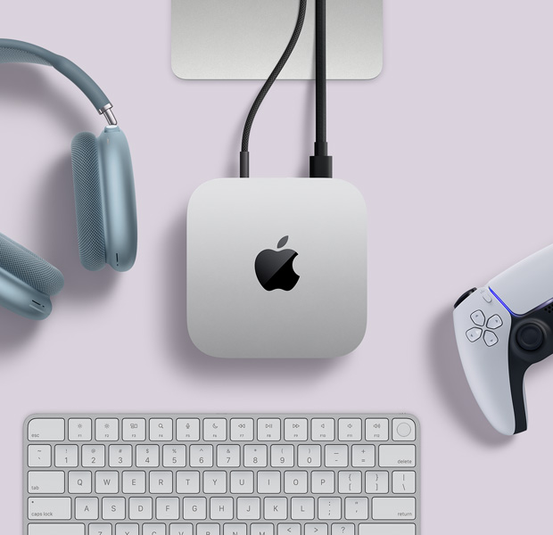 俯視圖展示銀色 Mac mini 擺放在桌上，有兩條連接線插在背面的連接埠，旁邊放有藍(lán)色 AirPods Max、一個(gè)銀色精妙鍵盤(pán)，以及一個(gè)無(wú)線遊戲手掣。