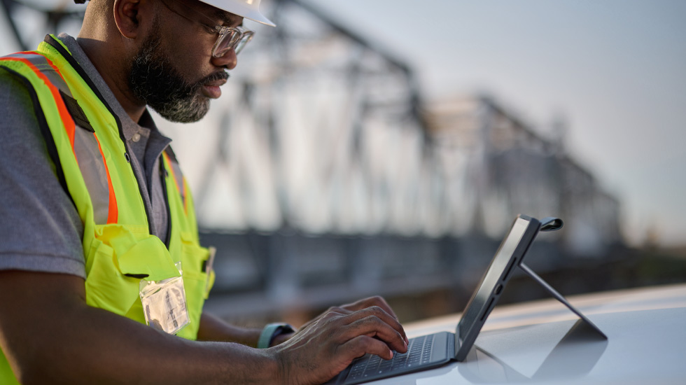 Un homme portant un casque de chantier tape sur le clavier d’un iPad. Il est en extérieur. On aper?oit un pont derrière lui.