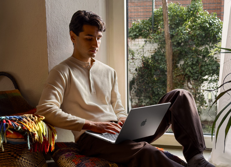 一個人坐在窗邊的地板上，使用 MacBook。