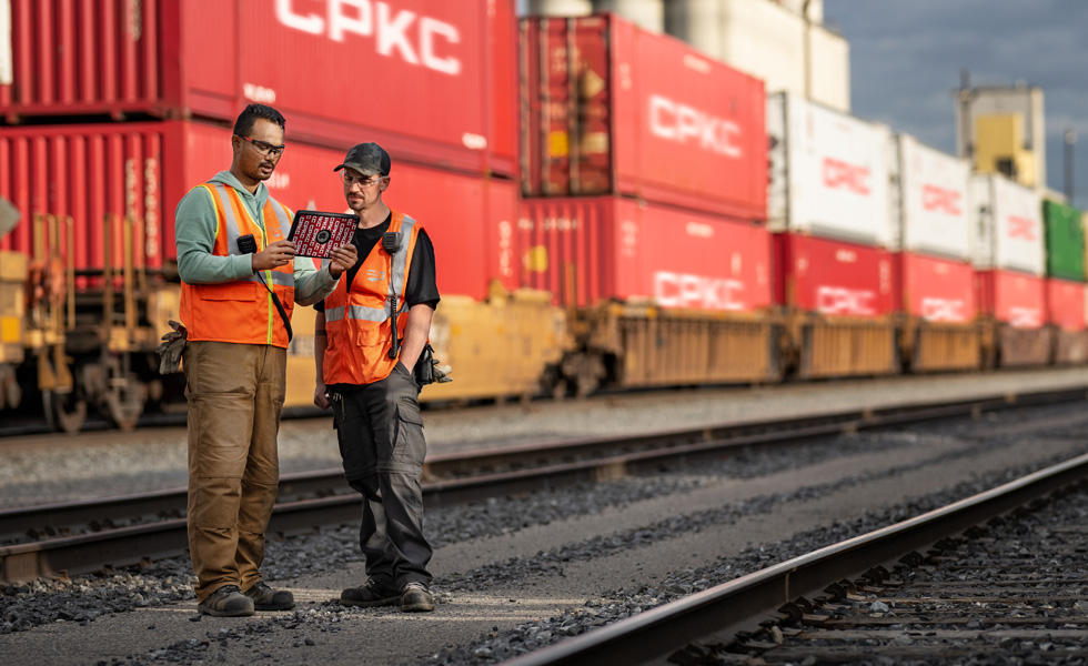 Zwei Bahnarbeiter blicken auf ein iPad, w?hrend sie auf einem Bahnhof neben einem stehenden Zug stehen, der Schiffscontainer transportiert