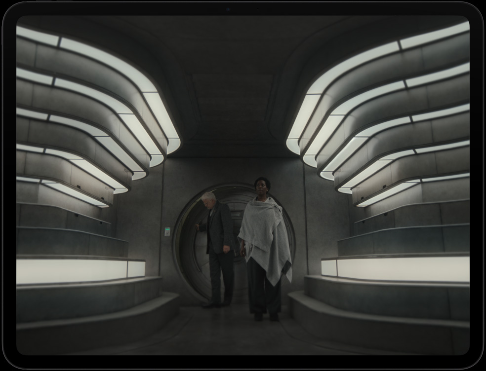 iPad Pro, parte delantera exterior, posición horizontal, color negro espacial, bisel de pantalla negro, la pantalla muestra una nave espacial futurista con maquinaria detallada y una persona con un traje espacial que interactúa con controles y equipos