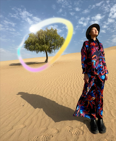 Imagen de una mujer con vestido multicolor de pie en un paisaje desértico con un solo árbol al fondo rodeado por un degradado arcoíris brillante que ilustra la capacidad de Apple Intelligence para borrar los objetos del fondo