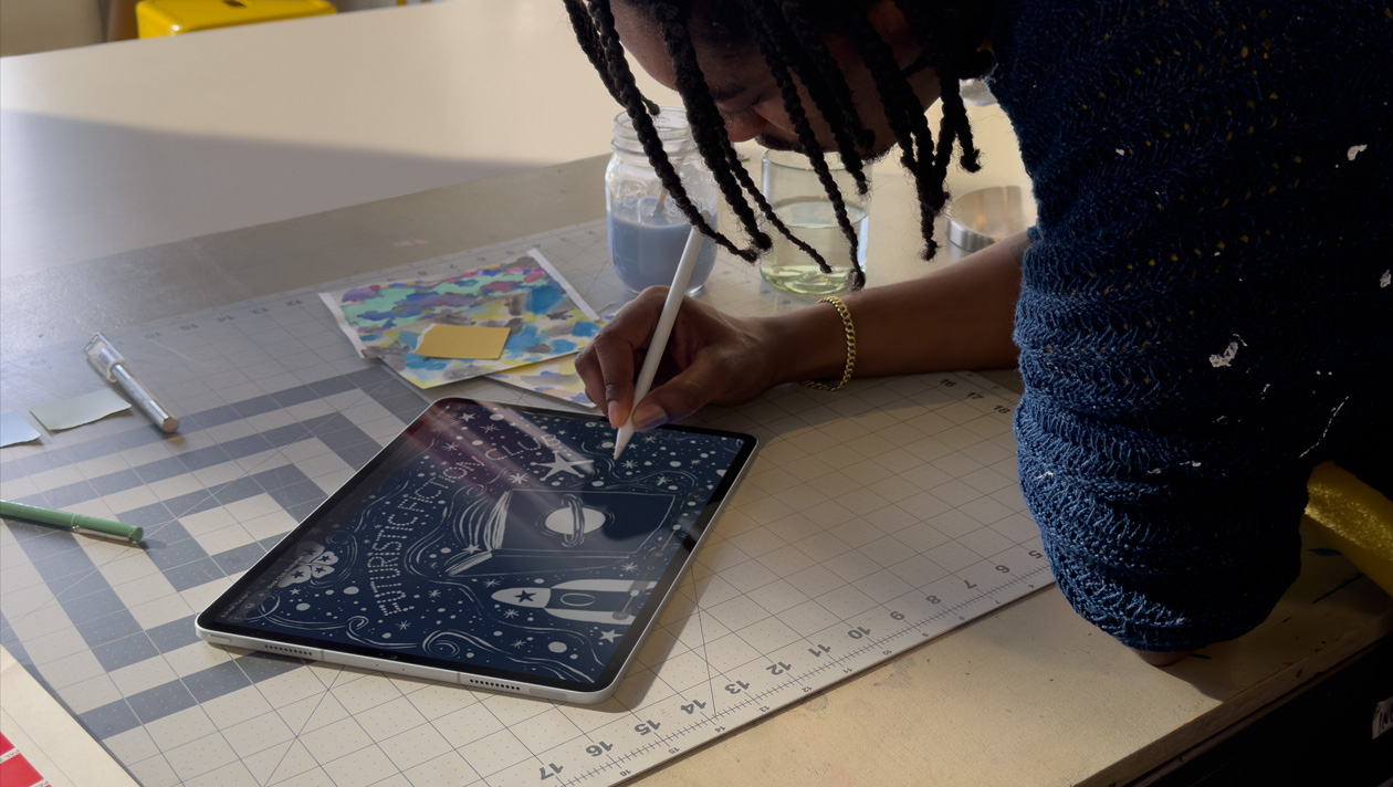 Una persona con trenzas y un suéter azul dibuja en un iPad Pro con un Apple Pencil, la pantalla muestra la ilustración de un cielo nocturno con un planeta en la portada de un libro, una nave espacial, una mariposa y la frase Futuristic Fiction Club escrita en el centro
